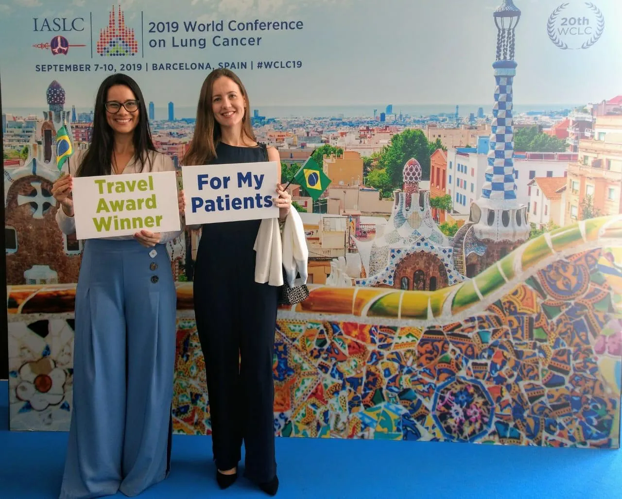 Gisele Fraga e Dra. Imanuely Borchardt diante do painel da 2019 World Conference on Lung Cancer, segurando placas com os dizeres Travel Award Winner e For My Patients