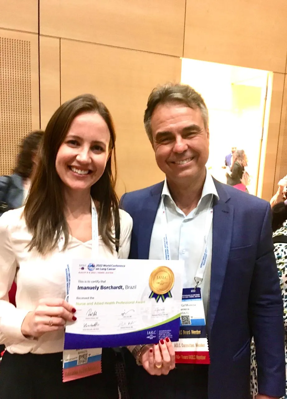 Dra. Imanuely Borchardt com o oncologista Dr. Carlos Gil Ferreira na WCLC 2022, em Viena, segurando o certificado do prêmio